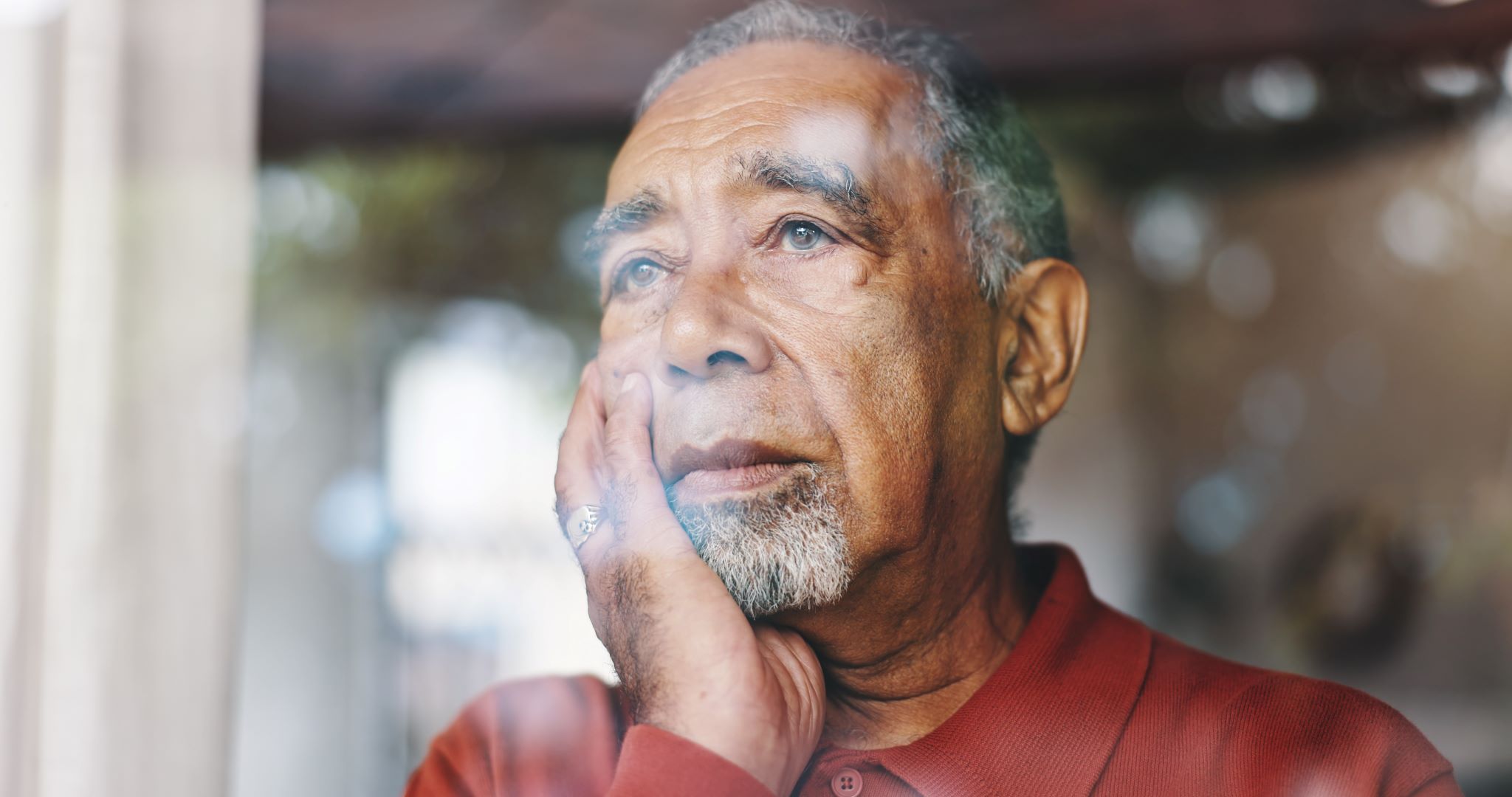 Senior man looking worried stares out the window of his apartment.